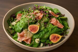 A bowl of fresh spinach topped with slices of prosciutto, figs, and microgreens drizzled with a light dressing.
