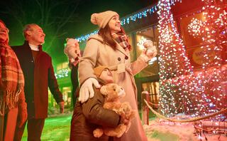 A joyful group enjoys a festive atmosphere, warmly dressed and admiring colorful holiday lights, with a child holding a teddy bear.