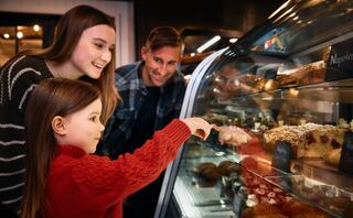 A young girl points excitedly at desserts in a bakery, accompanied by a smiling woman and a man, enjoying a delightful moment together.
