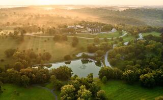 A serene landscape at sunrise features rolling hills, lush greenery, a calm pond, and a distant building, bathed in soft, golden light.