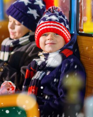 Two cheerful boys in winter attire, one in a colorful knit hat and scarf, sit indoors, enjoying a festive atmosphere and playful snowflakes.