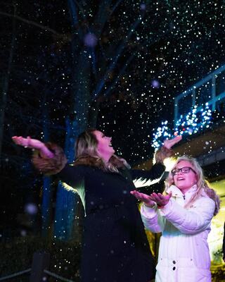 Two women joyfully catch falling snowflakes at night, surrounded by twinkling lights and a festive atmosphere.