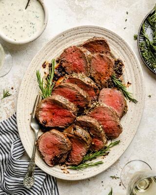 Sliced roast beef served on a platter, garnished with fresh herbs, alongside a creamy sauce and accompanied by glasses of white wine.