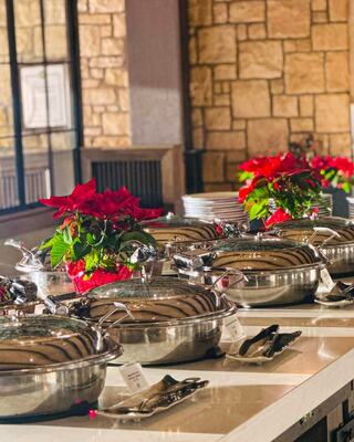 A buffet spread showcases silver serving dishes with lids, surrounded by festive red poinsettias, set against a warm stone wall.