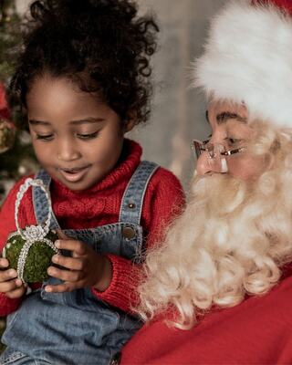 A joyful child in festive attire admires a Christmas ornament while sitting next to a jolly Santa Claus, surrounded by holiday decor.