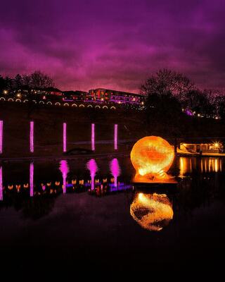 A glowing orange sphere reflects in water, surrounded by purple skies and illuminated structures along the shore, creating a serene atmosphere.