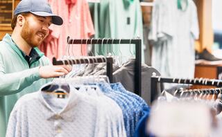A man browses through clothing racks in a store, smiling as he examines various shirts. Items in pastel colors hang in the background.