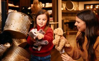 A young girl and a woman smile as they explore plush toys, surrounded by a cozy store setting filled with warm, inviting decor.