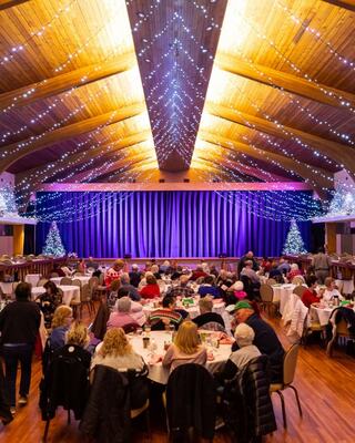 A festive venue with elegant lighting, tables filled with guests, and a stage adorned with purple drapes and holiday decorations.