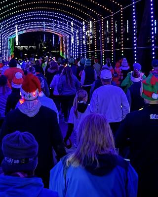 A festive crowd walks through a brightly lit tunnel adorned with colorful lights, wearing holiday hats and cheerful attire at Oglebay's Twinkle Trot 5K.