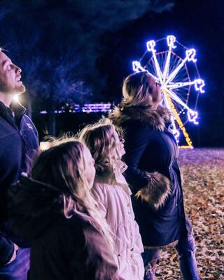 A family stands together, gazing up in awe at a brightly lit ferris wheel against a night sky. Colorful lights create a festive atmosphere.