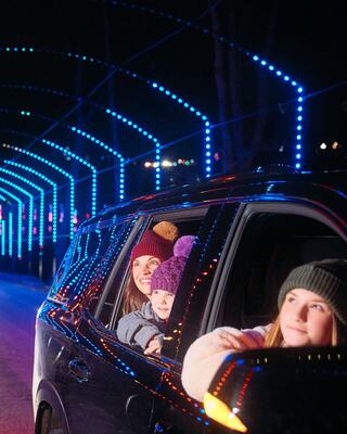 Three smiling individuals lean out of a car window, surrounded by a vibrant display of colorful lights in a nighttime setting.