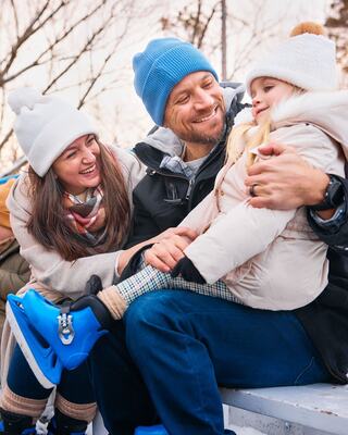 A happy family shares a joyful moment outdoors, with a child in skates being playfully embraced by parents, all bundled in winter clothing.
