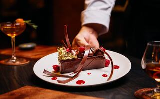 A gourmet dessert featuring rich chocolate, garnished with raspberries, elegant chocolate curls, and a touch of gold, accompanied by cocktails.