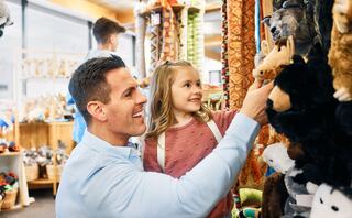 A man and a young girl smile while browsing a store filled with colorful stuffed animals and toys, enjoying a fun shopping moment together.