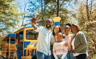 A happy family takes a selfie together in a scenic outdoor setting, with a colorful train in the background. Everyone smiles brightly.