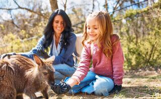 A woman and a girl joyfully interact with a small animal in a sunny outdoor setting, showcasing a moment of connection and care.