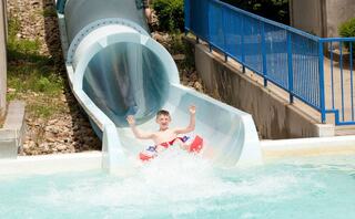 A joyful child slides down a water slide, splashing into a pool, with arms raised in excitement. Bright, sunny day with green surroundings.