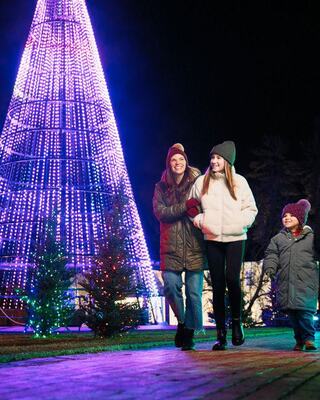 Three people walk together at night, adorned in winter wear, with a brightly lit Christmas tree and festive decorations in the background.