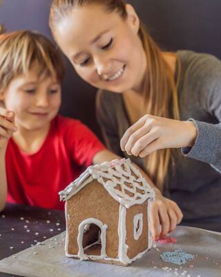 A woman and a young boy joyfully decorate a gingerbread house with icing and colorful sprinkles, sharing a festive moment together.