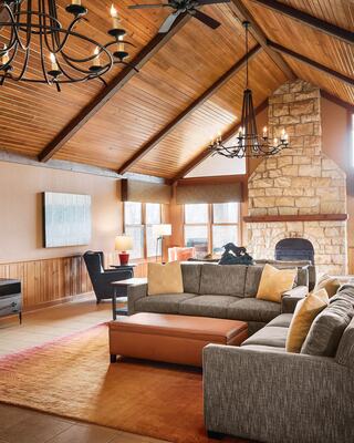 A cozy living room features a high wooden ceiling, stone fireplace, modern couch with cushions, and large windows bringing in natural light.