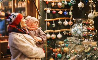 A woman holds a child, both gazing in wonder at colorful holiday ornaments displayed in a festive shop adorned with twinkling lights.