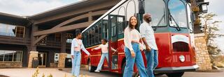 A cheerful family disembarks from a red trolley outside a rustic building, enjoying a fun outing together. Bright smiles and casual outfits abound.