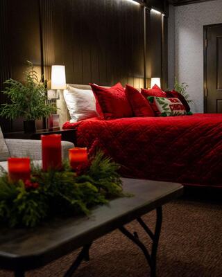A cozy bedroom decorated in red and green hues, featuring a plush bed, warm lighting, and festive accents on a table.