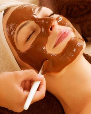 A woman relaxes with a chocolate face mask applied, eyes closed in tranquility, while someone gently helps with the application.