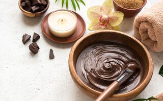 A wooden bowl of smooth chocolate mixture, surrounded by chocolate pieces, a candle, sugar, a towel, and a delicate orchid.