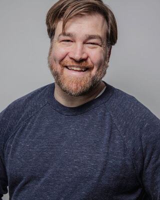 A smiling man with a beard wears a navy sweater, exuding warmth and friendliness against a plain background.