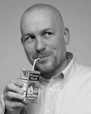 A bald man playfully sips from a juice box, featuring a mischievous smile, embodying a lighthearted and youthful spirit.
