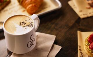 A latte topped with lavender sits on a wooden table beside a croissant and a pastry decorated with raspberries and a golden crust.