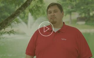 A man in a red polo shirt stands near a tranquil pond, surrounded by greenery, with a fountain visible in the background.