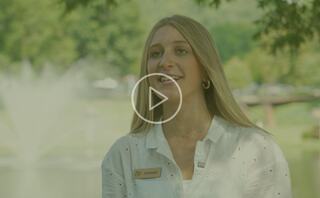 A young woman speaks in front of a scenic backdrop with a fountain, sharing insights while wearing a name tag and casual attire.