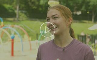 A young girl with long hair smiles while speaking, with a colorful playground in the background, creating a cheerful atmosphere.