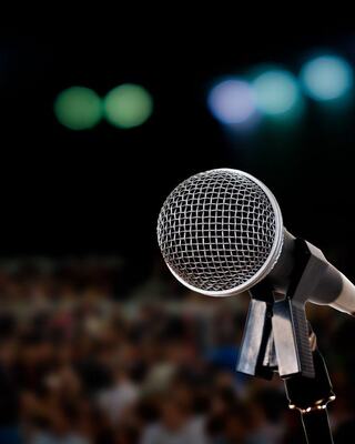 A close-up of a microphone on a stand, with blurred colorful lights and an audience in the background, creating an atmosphere for performance.