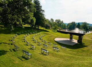 A landscaped outdoor amphitheater features a circular arrangement of benches facing a stage near a serene pond, surrounded by lush greenery.