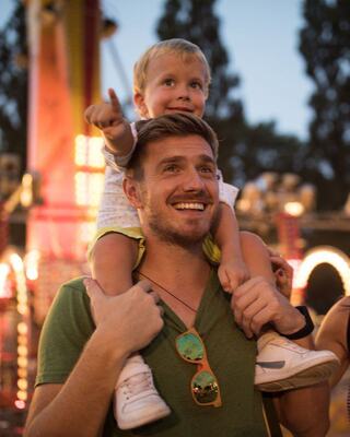 A smiling man carries a young boy on his shoulders, both looking excited as they enjoy a lively, brightly lit fairground atmosphere.