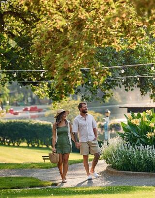 A couple strolls hand in hand through a lush garden, surrounded by greenery and flowers, enjoying a sunny day.