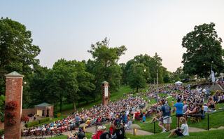 A large crowd gathers on a hillside, surrounded by trees, enjoying an outdoor event as the sun sets in the background.