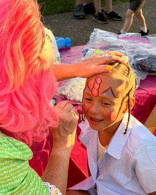 A child smiles as a performer with pink hair paints colorful designs on their face at a festive outdoor event.