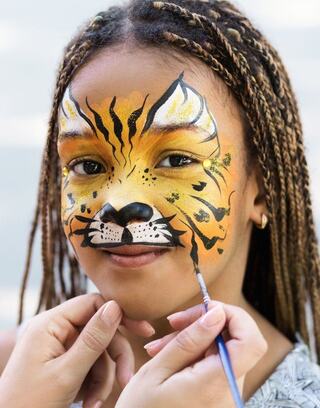 A girl has her face painted as a tiger, displaying vibrant orange, black, and white designs, with a joyful expression.