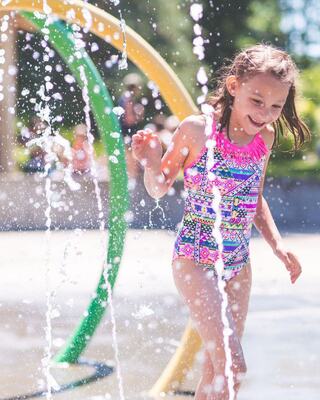 A joyful girl in a colorful swimsuit plays in a water spray, smiling as droplets scatter around her on a sunny day.