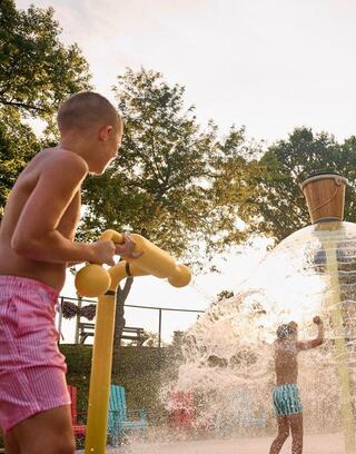 Two boys play at a water park, joyfully splashing in the sunlight. One boy is holding a water sprayer while the other dances in the spray.