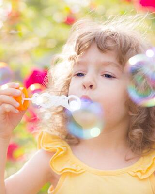 A young girl with curly hair blows bubbles, wearing a sunny yellow outfit, surrounded by colorful blooms in a bright and cheerful setting.