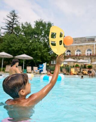 A child plays in a pool, joyfully reaching for a colorful ball with a yellow scoop, surrounded by other swimmers and sunny greenery.