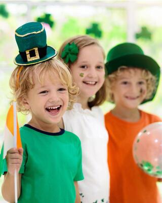 Three smiling children celebrate with festive hats and colorful attire, surrounded by St. Patrick's Day decorations.