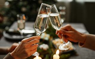 Two hands clink champagne flutes together in a festive setting, with blurred glasses and soft candlelight creating a warm atmosphere.