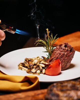 A gourmet plate features a beautifully cooked beef cut, accompanied by grilled vegetables and a sprig of rosemary, with a torch adding final touches.